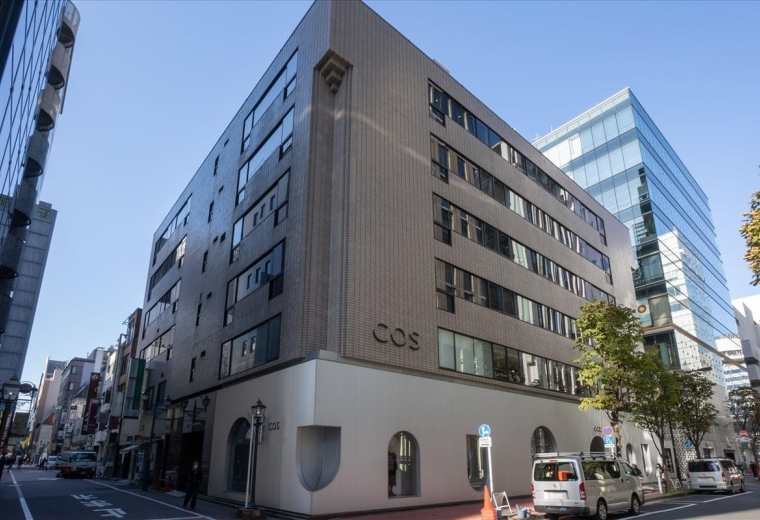 Corner exterior view of the Okura Bekkan building featuring a modern tan facade and street-level shopfronts.