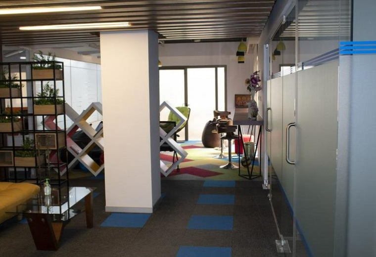 Spacious communal lounge at Indiqube Ramky with modern shelving, colourful rugs, and a frosted glass partition.