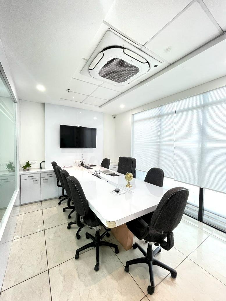 Spacious boardroom at Atulya IT Park with a long white table, black chairs, and large windows with blinds.