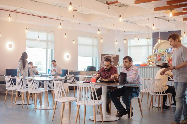 Spacious communal dining area and cafe at Global Business Park Tower D with white chairs and hanging bulb lights.
