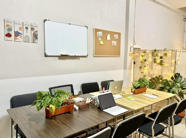 Long wooden meeting table with laptops, plants, and whiteboards in a bright workspace.