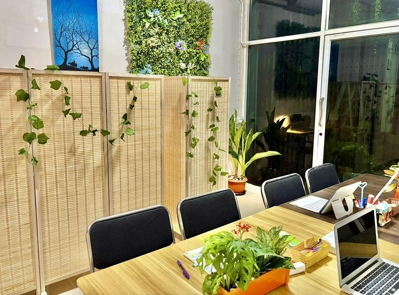 Shared workspace with natural wood tables, black chairs, and abundant indoor plants.