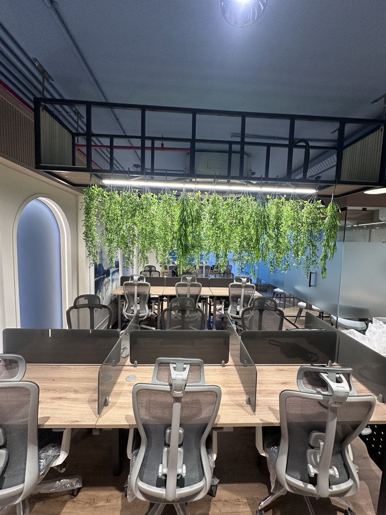 Open-plan office with wooden desks and lush artificial greenery hanging from the ceiling.