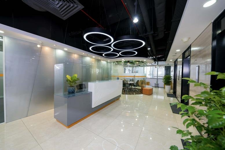 Spacious office reception area with a minimalist white desk, circular ceiling lights, and indoor greenery.