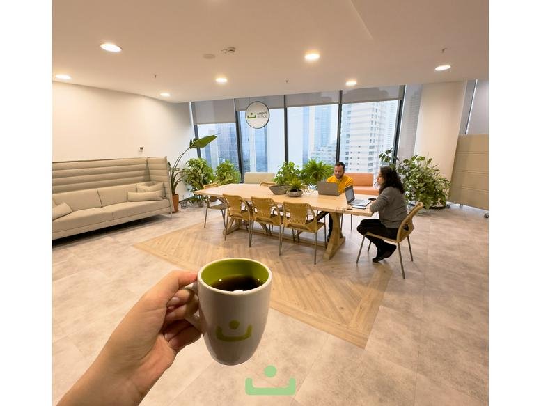 Bright communal lounge with a wooden table, indoor plants, and a person enjoying coffee.