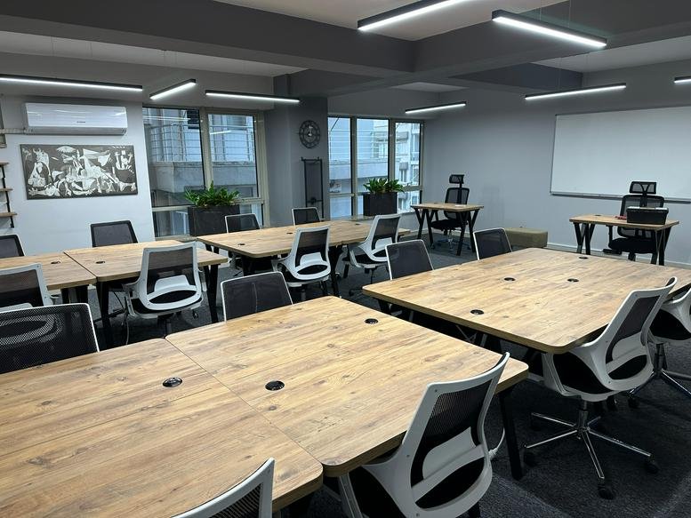 Spacious open-plan coworking area with long wooden tables, white chairs, and bright overhead lighting.