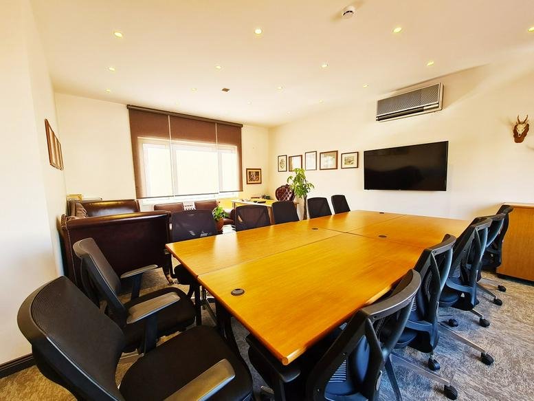 Spacious boardroom with a long wooden table, black mesh chairs, and a large wall-mounted monitor.