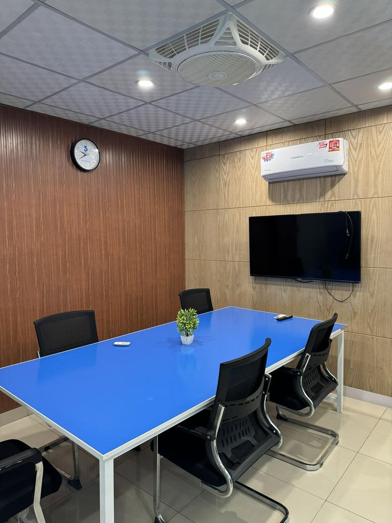 Meeting room with a bright blue table, black mesh chairs, and a wall-mounted TV screen.