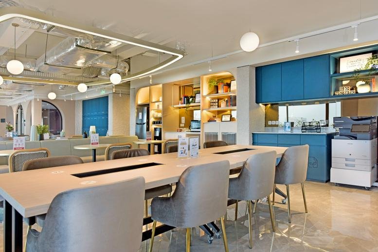 Long wooden collaboration desk with grey chairs in a sunlit open-plan office space.