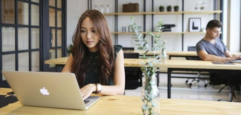 Coworking space at The Hive Prakanong, Bangkok with people working at wooden desks by a plant.