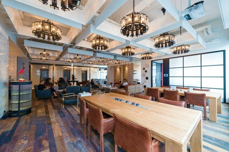 Spacious coworking lounge with wooden communal tables, leather chairs, and industrial chandeliers.