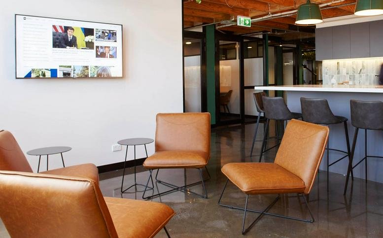 Breakout lounge with tan leather chairs and a wall-mounted digital screen near a kitchen counter.