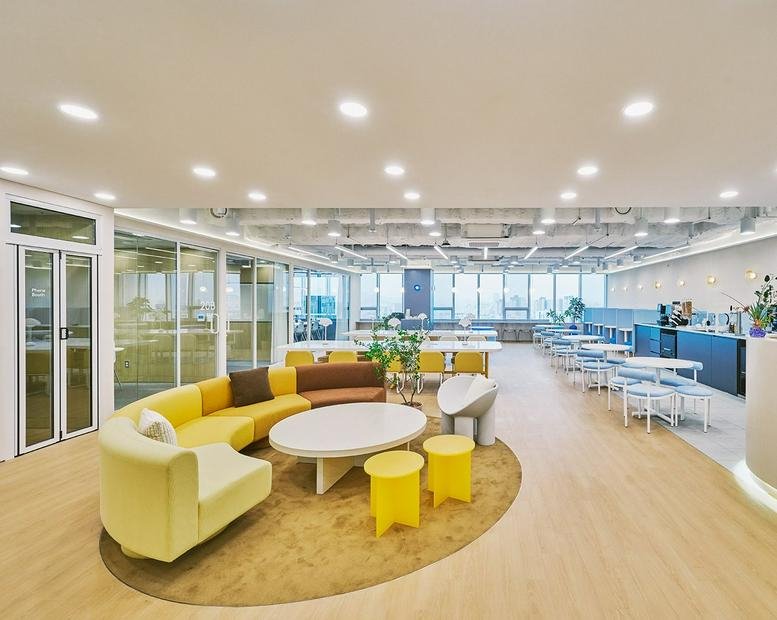 Spacious coworking lounge with a curved yellow sofa, round coffee table, and wood-look flooring.
