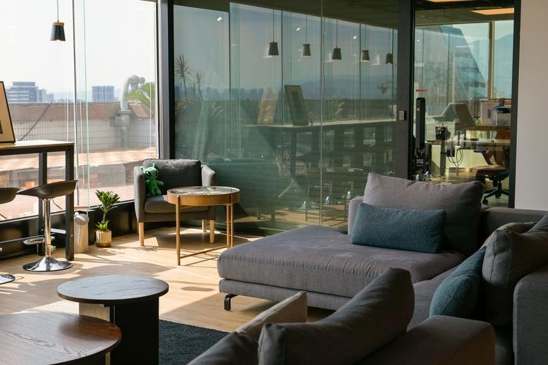 Sunny lounge area with grey sofa, wooden tables, and floor-to-ceiling windows.