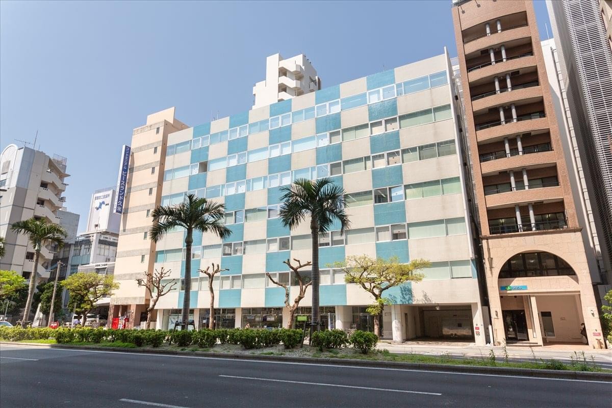 The checkered blue and white exterior of the 5F COI Naha Building with palm trees out front.