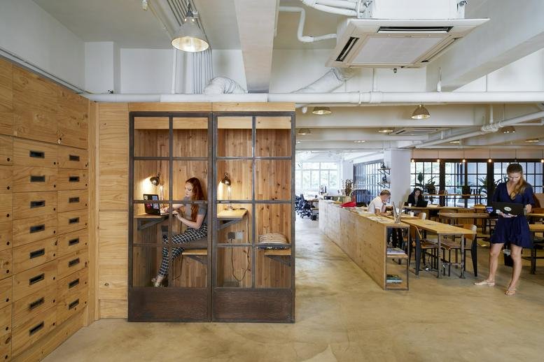 Spacious open-plan workspace with wooden desks, a glass-walled booth, and high ceilings.