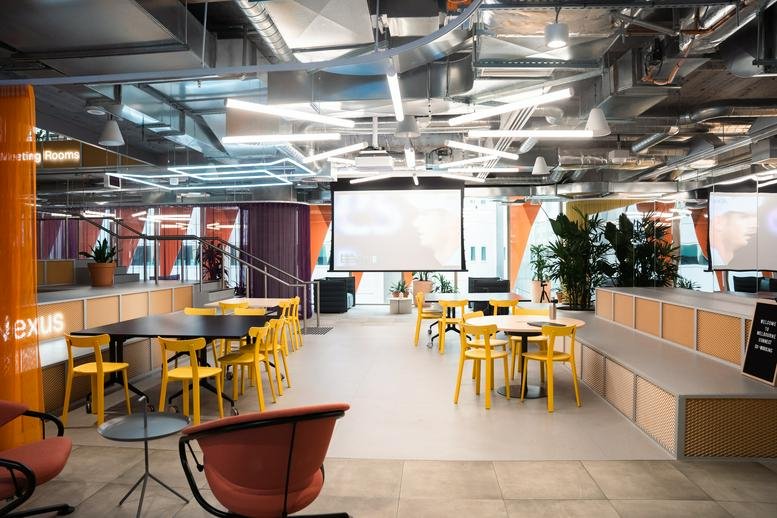 Large breakout space with yellow seating, a projector screen, and indoor greenery.