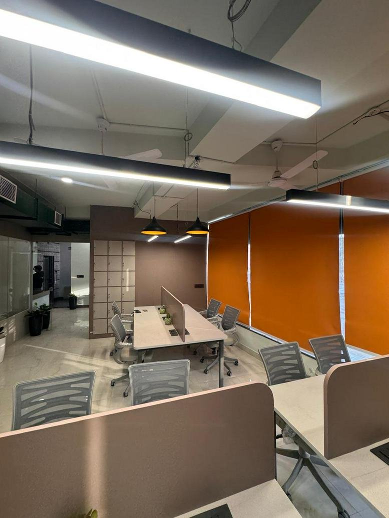Open-plan office area featuring long desks, orange window blinds, and linear pendant lighting.