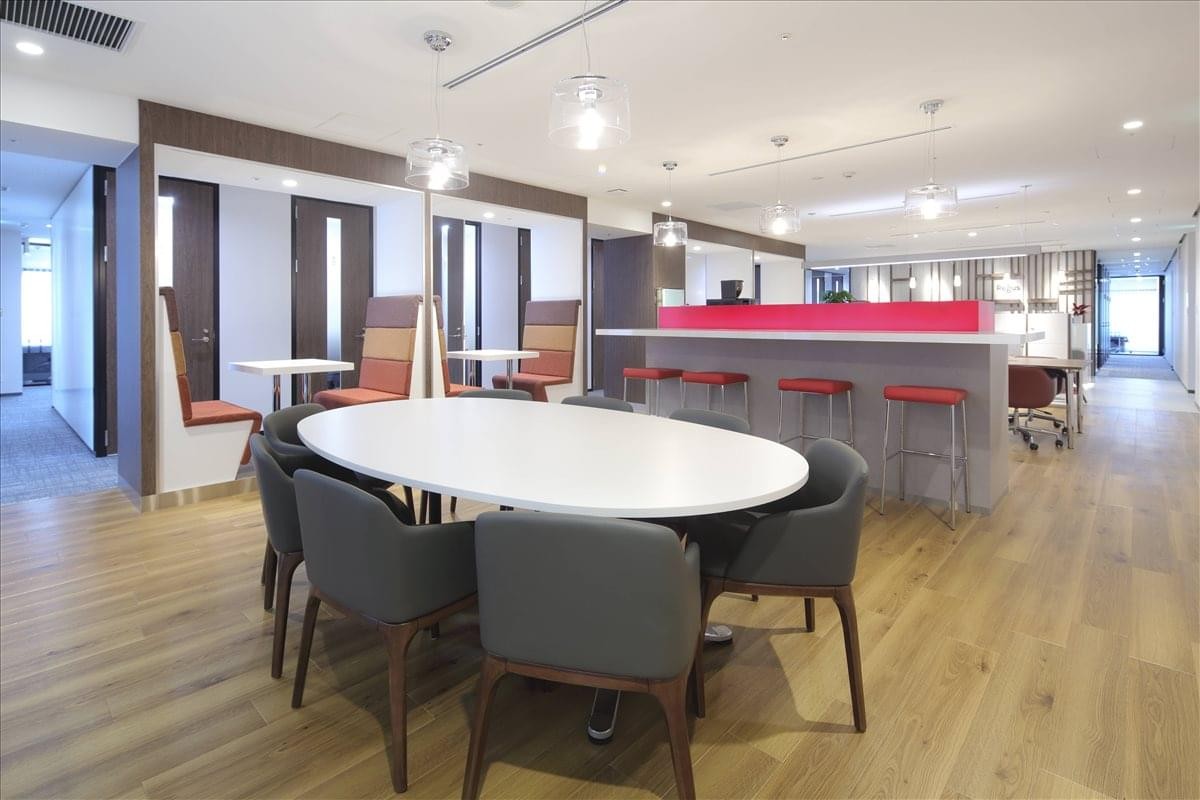 Modern communal breakout area with a large oval white table, grey chairs, and a bar counter with red stools.