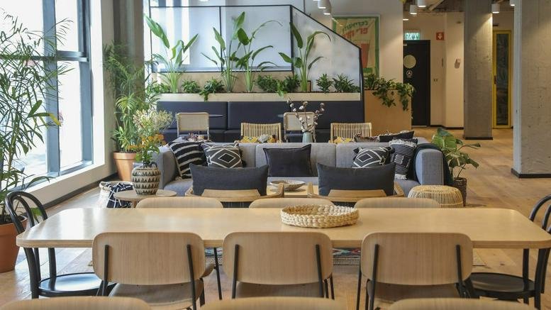 Spacious communal lounge at 23 Schocken Street with plush sofas, wooden tables, and lush indoor plants.