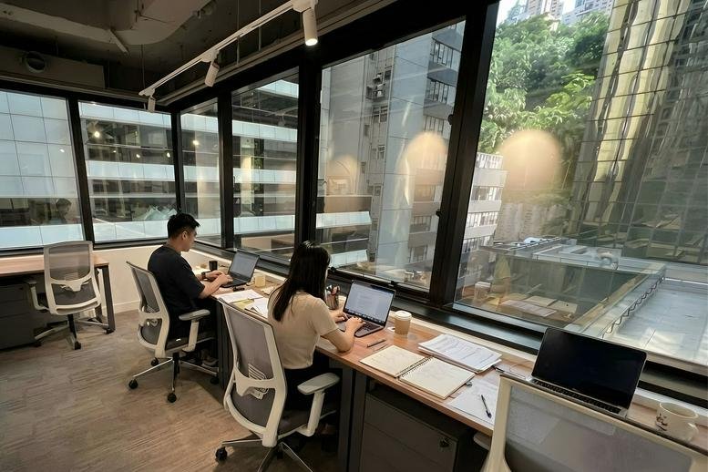Workspace at Ruttonjee Centre Dina House with long desks facing large windows overlooking the city.