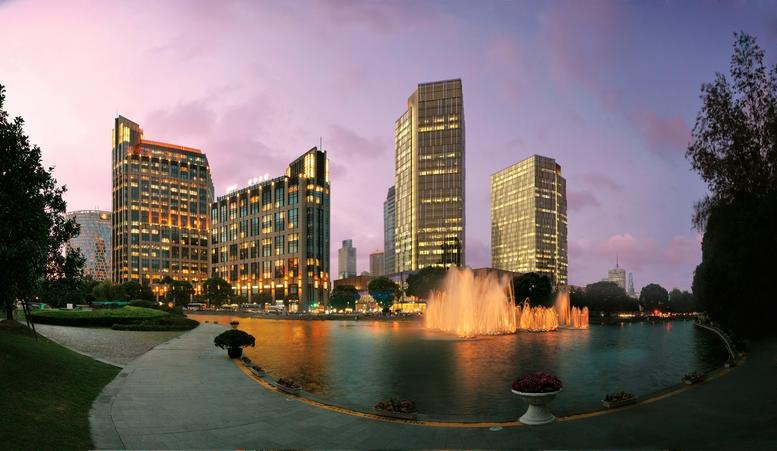 City skyline at dusk featuring illuminated buildings and a colorful fountain in a central park.
