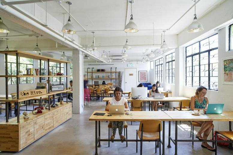 Bright open-plan coworking space with people working at long wooden tables.