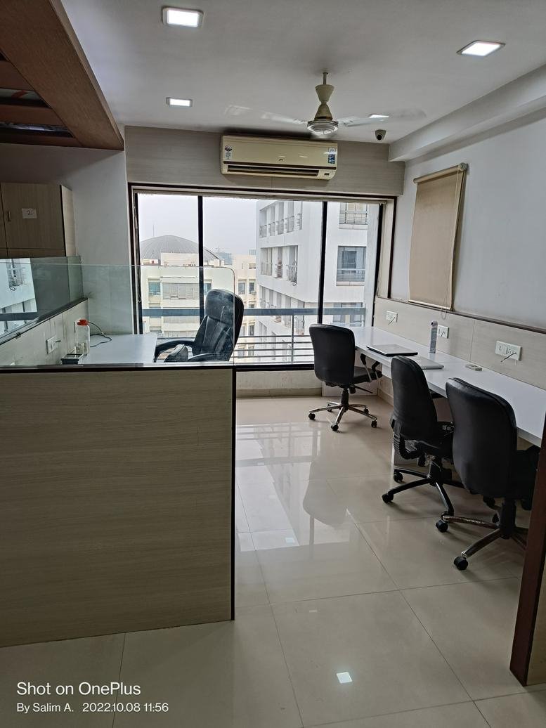 Open-plan office with multiple workstations, black ergonomic chairs, and large windows.