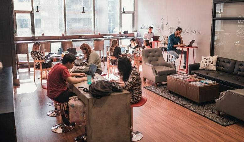 Lively coworking space with people working at wooden tables and a plush grey armchair.