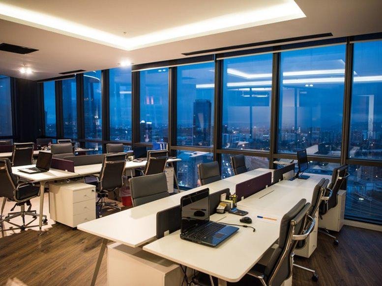 Modern workspace featuring long white desks and floor-to-ceiling windows with city evening views.