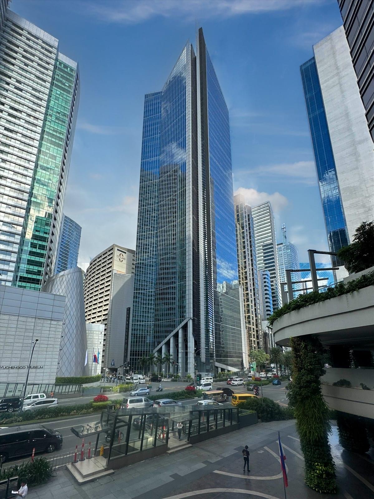 View of the towering glass skyscraper from a street-level park area with lush greenery.