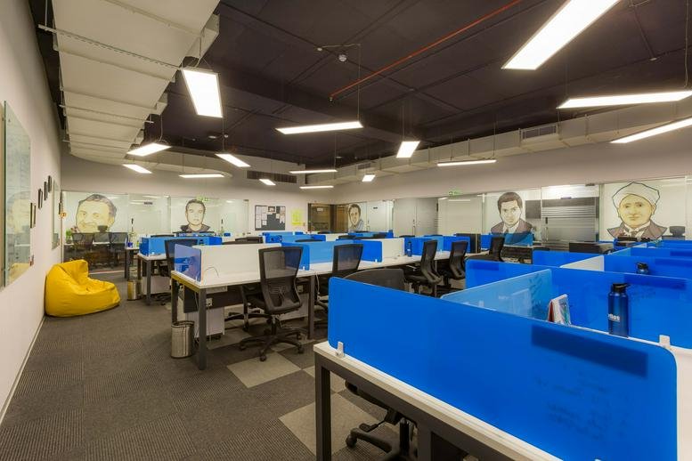 Large open-plan office with blue partitioned desks and illustrated portraits on the walls.