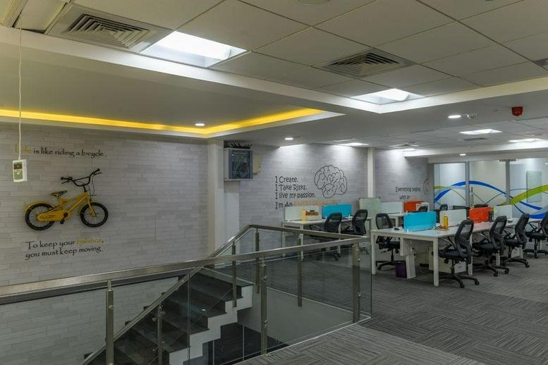 Spacious communal lounge and workspace at IndiQube Delta with a decorative wall bicycle and modern staircase.