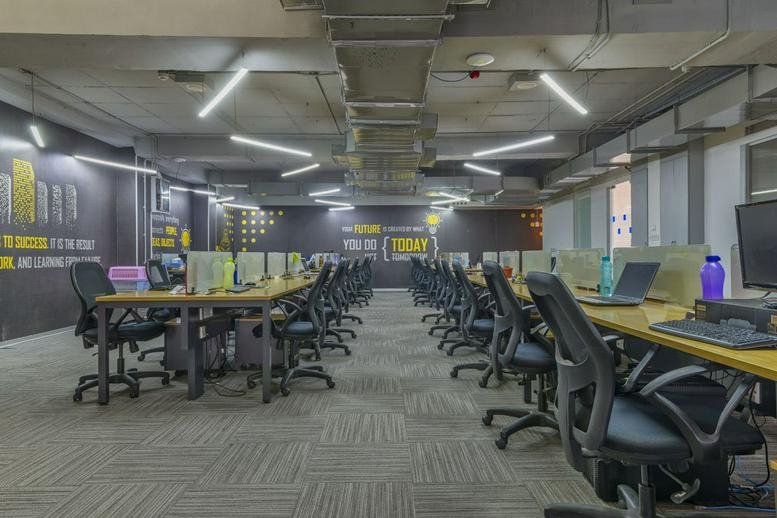 Spacious open-plan coworking area at IndiQube ETA with long wooden desks and ergonomic black chairs.