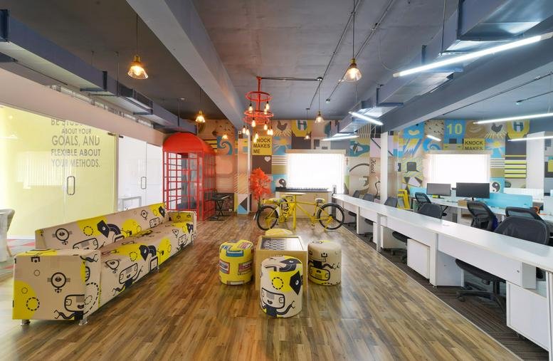 Colorful communal lounge at IndiQube Gamma with a long sofa, artistic stools, and wooden flooring.