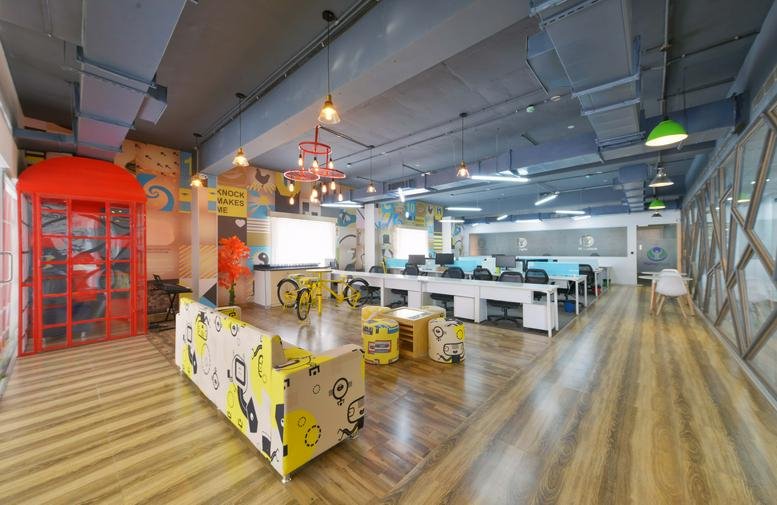 Spacious coworking lounge at IndiQube Orion with colorful seating, wood floors, and a red telephone booth.