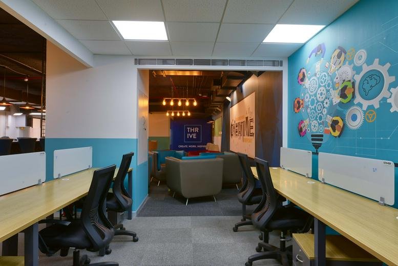 Open-plan workspace at Indiqube The Leela, Old Airport Road featuring wooden desks and a colorful graphic feature wall.