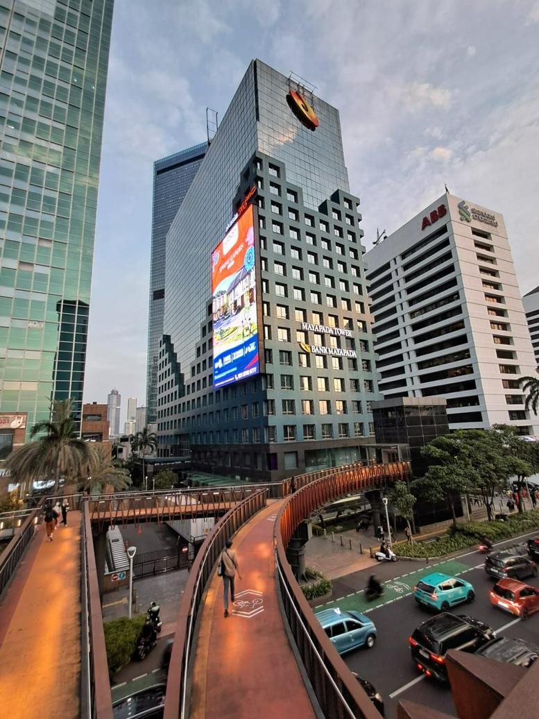 A modern pedestrian bridge curves toward Mayapada Tower in a busy urban setting.
