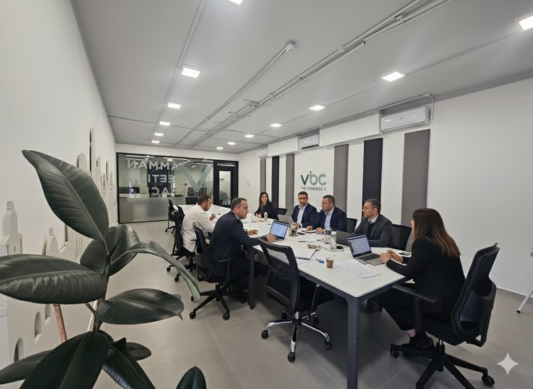 Large meeting room with professionals seated around a conference table with laptops and a green plant.