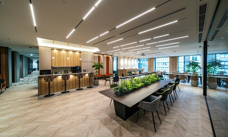 Vibrant communal area with a cafe-style bar, indoor plants, and a long wooden table under bright linear lighting.