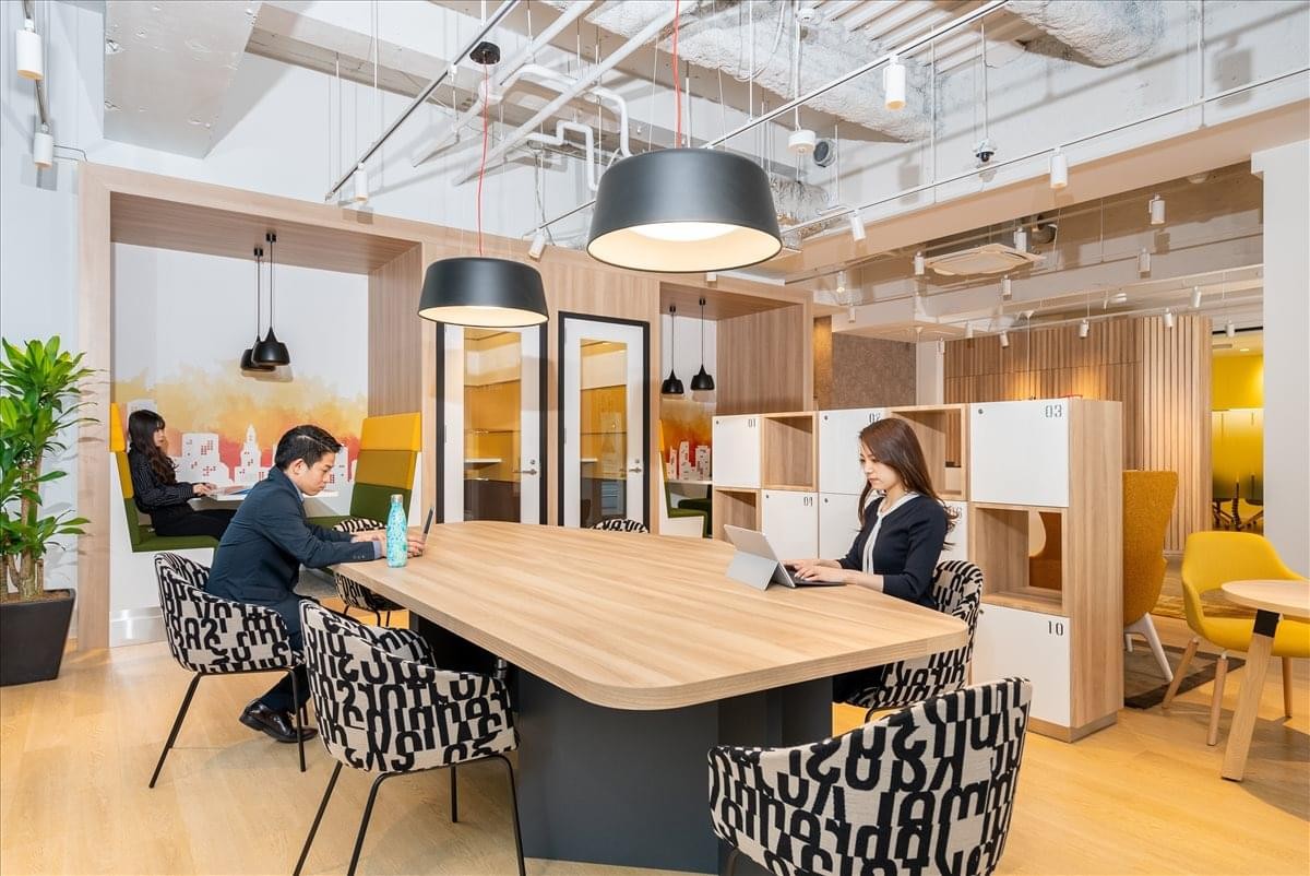 Modern coworking area with people working at a large wooden table under industrial lighting.