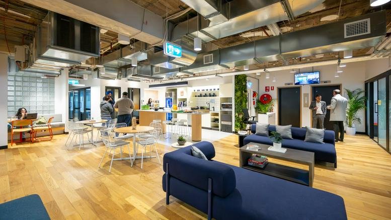 Spacious breakout area with blue sectional sofas, industrial ceilings, and people socializing.